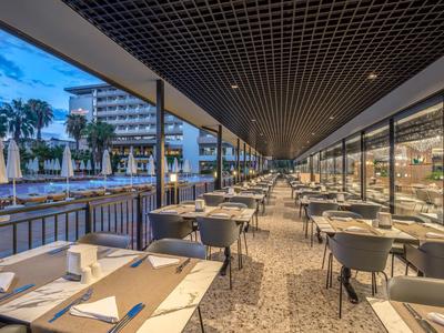 Terraza moderna de hotel con mesas, sillas y vista a la piscina al anochecer.