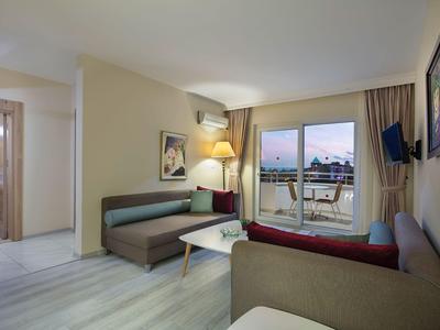 Sala moderna de hotel con sofá, mesa, televisor y balcón con vista al mar al atardecer.
