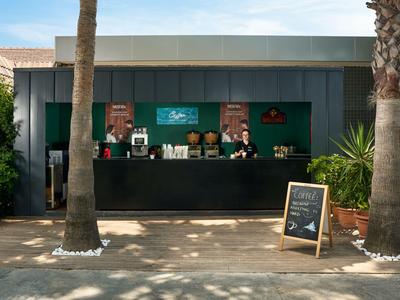 Moderno quiosco de café al aire libre entre palmeras con terraza y menú del día.