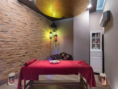 Ruheraum mit Massagetisch, rotem Tuch, Stehlampe, Steinwand und goldenem Deckendekor.
