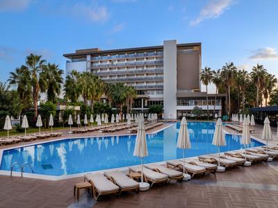Complejo hotelero moderno con gran piscina, tumbonas y palmeras bajo un cielo azul.