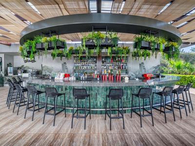 Bar semicircular de hotel con mostrador verde, taburetes negros y plantas decorativas.