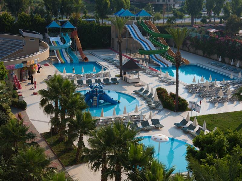 Hotelzwembad met glijbanen, ligstoelen en palmbomen in een zonnige omgeving.