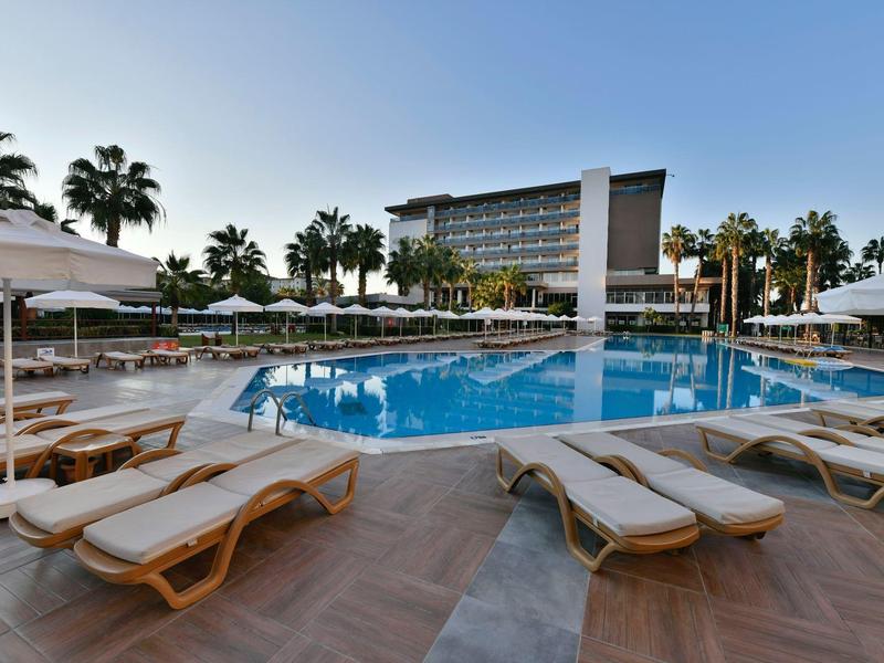 Groot buitenzwembad met ligbedden en parasols naast een modern hotelgebouw onder een heldere hemel.