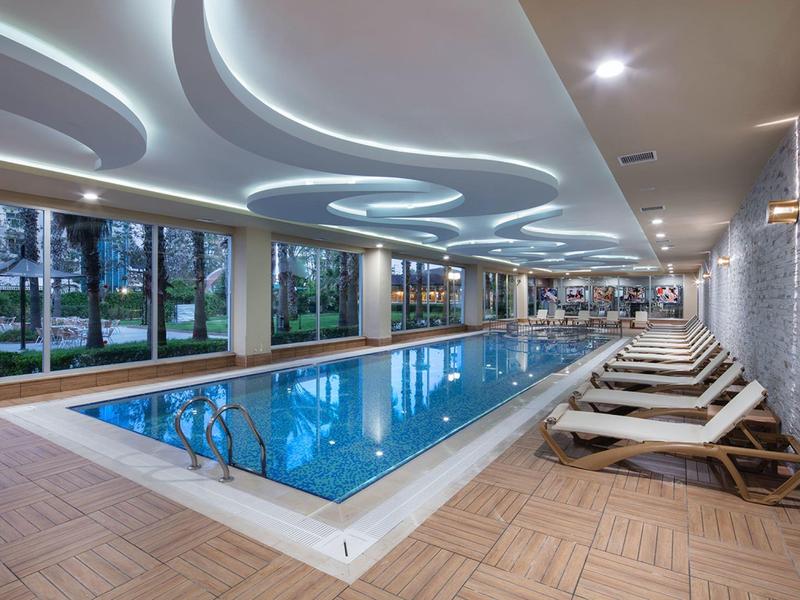 Licht overdekt zwembad met ligstoelen naast het water en grote ramen met uitzicht op een park.