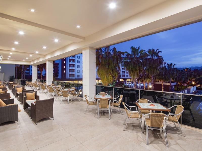 Groot hotelterras met tafels en stoelen, verlicht en met uitzicht op palmbomen bij schemering.