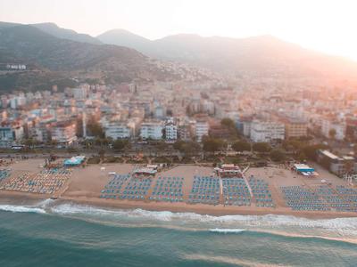 Strand mit Liegen und Sonnenschirmen vor einer Stadt am Fuße bewaldeter Hügel bei Sonnenuntergang.