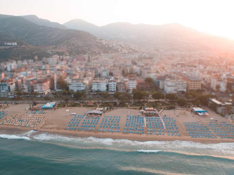 Strand mit Liegen und Sonnenschirmen vor einer Stadt am Fuße bewaldeter Hügel bei Sonnenuntergang.