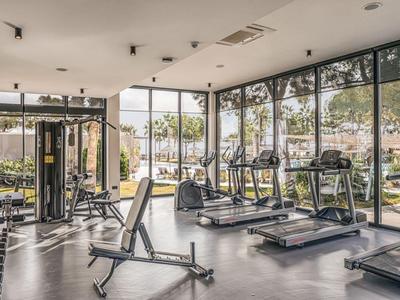 Salle de sport lumineuse avec tapis de course, poids et grandes fenêtres donnant sur un parc.