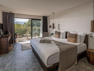 Moderní hotelový pokoj s velkou postelí, posezením a výhledem na terasu s přírodní scenérií.