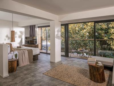 Světly hotelový pokoj s velkými okny, kobercem a výhledem na zahradu.