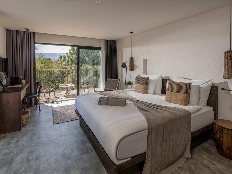 Moderní hotelový pokoj s velkou postelí, posezením a výhledem na terasu s přírodní scenérií.