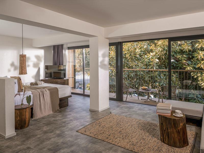 Světly hotelový pokoj s velkými okny, kobercem a výhledem na zahradu.