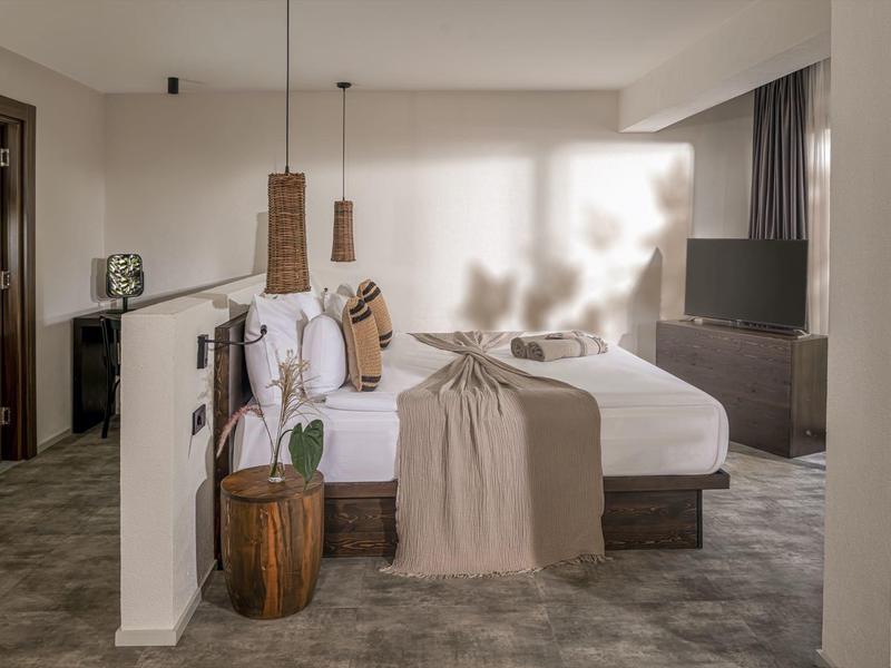 Moderní hotelový pokoj s manželskou postelí, kobercem a minimalistickým zařízením v neutrálních tónech.