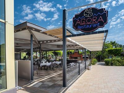 Außenbereich eines Restaurants mit weißen Tischen und Stühlen unter einem Sonnendach bei blauem Himmel.