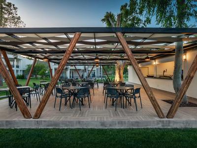 Offener Pavillon mit Holzbalken, Tischen und Stühlen, umgeben von grünem Garten.