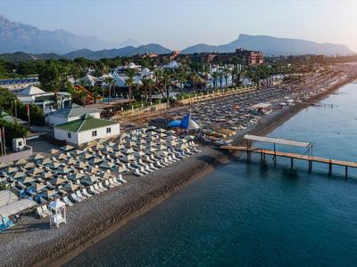 Langer Sandstrand mit Sonnenschirmen, Liegen und Steg, dahinter zahlreiche Hotelanlagen vor Bergen.