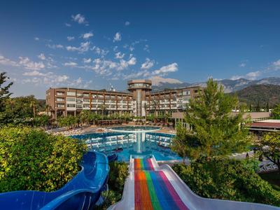 Hotel mit großem Außenpool, bunten Wasserrutschen und bewaldeter Umgebung unter blauem Himmel.