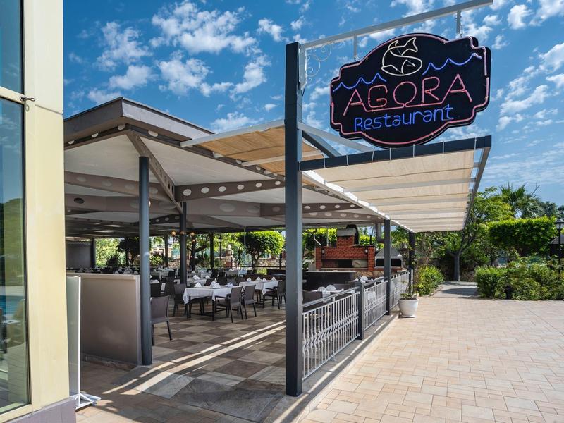 Außenbereich eines Restaurants mit weißen Tischen und Stühlen unter einem Sonnendach bei blauem Himmel.