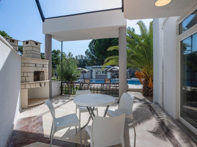 Terrasse mit weißem Tisch, Stühlen, gemauertem Grill und Blick auf Pool und Bäume im Hintergrund.