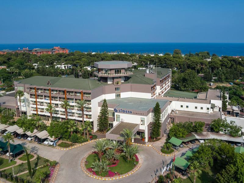 Großes Hotelgebäude mit Pool, umgeben von grünen Bäumen und nah am blauen Meer.