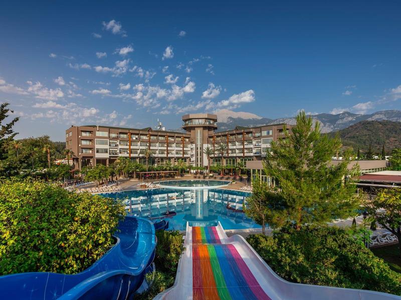 Hotel mit großem Außenpool, bunten Wasserrutschen und bewaldeter Umgebung unter blauem Himmel.