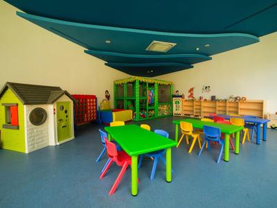 Bunter Kindergartenraum mit kleinen Tischen, Stühlen, Spielhaus und Aufbewahrungsschrank.
