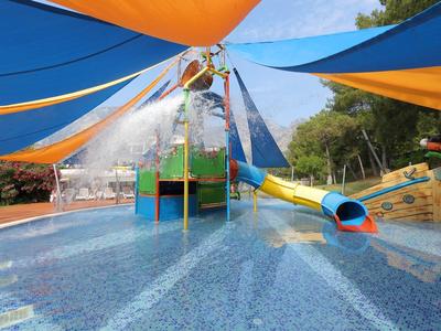 Bunter Wasserspielplatz mit Schirmdächern, Rutsche und Wasserspray, umgeben von Bäumen.