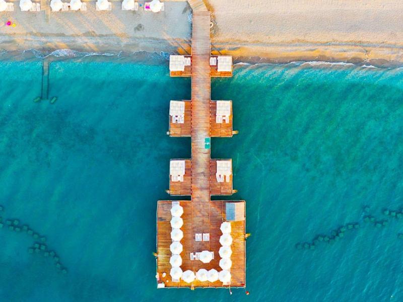 Luchtfoto van een houten pier in helder turquoise zee naast een zandstrand met ligbedden.