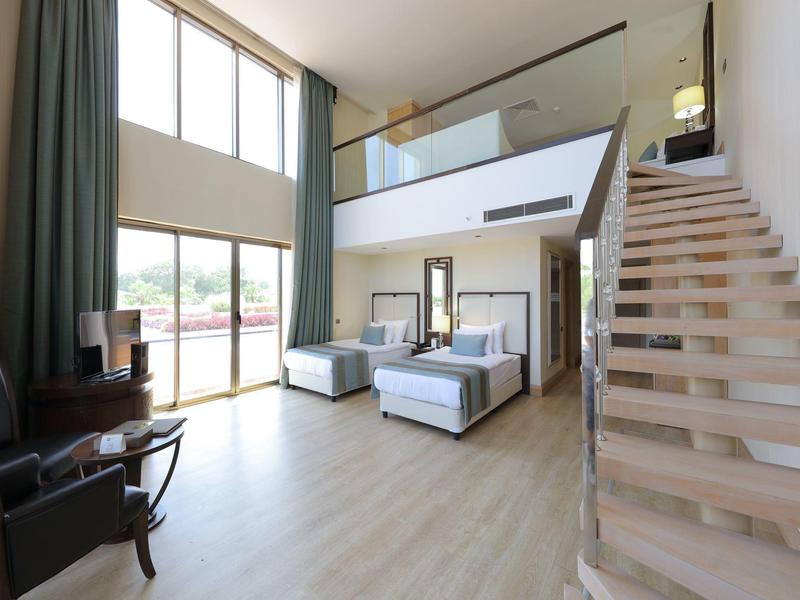 Ruime hotelkamer met twee eenpersoonsbedden, grote ramen, houten trap en stijlvolle zithoek.