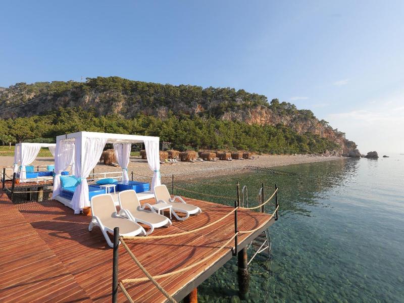 Houten pier met cabana's en ligbedden met uitzicht op de kalme zee en een rotsachtige heuvel op de achtergrond.