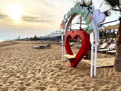 Cadre de balançoire en forme de cœur rouge sur plage de sable avec chaises longues et coucher de soleil.
