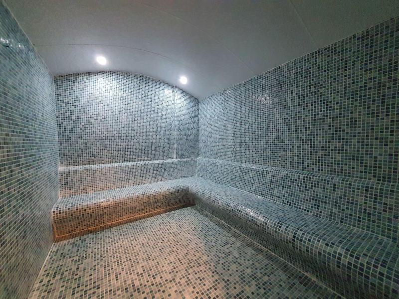 Binnen sauna met mozaïektegels en ingebouwde bank, verlicht door plafondlampen.
