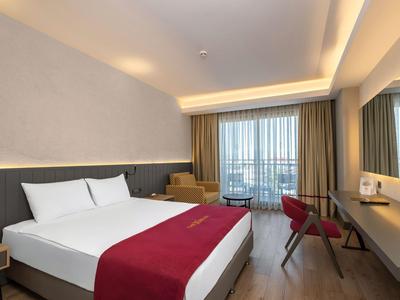 Camera d'hotel moderna con letto matrimoniale, copriletto rosso e balcone con vista sulla città.