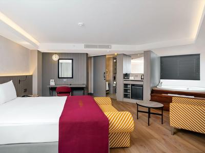 Camera d'hotel moderna con letto matrimoniale, scrivania, zona salotto e angolo cottura.
