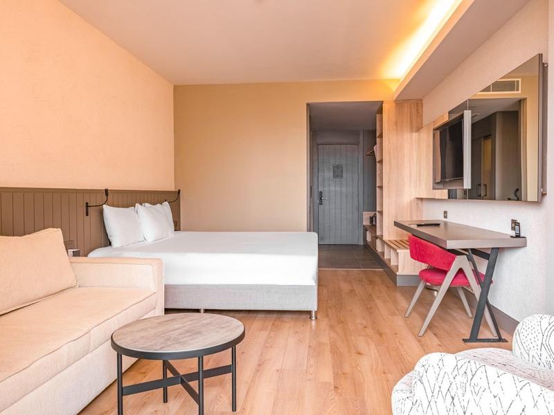 Habitación de hotel luminosa con cama, sofá, mesa pequeña y escritorio con silla.