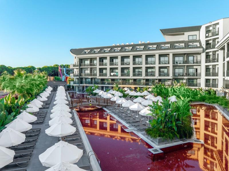 Complejo hotelero moderno con piscina, sombrillas y tumbonas rodeado de paisaje verde.