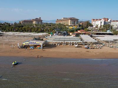 Pláž s restauracemi a hotely v pozadí, klidné moře, písčitá pláž v popředí.
