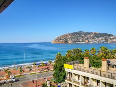 Vue sur la mer et une île, avec des palmiers et des bâtiments au premier plan sous un ciel bleu.
