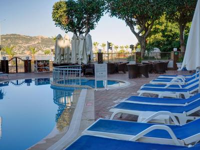 Pool mit blauer Liegefläche, weißen Sonnenschirmen und Bäumen, im Hintergrund Hügel mit Gebäuden.