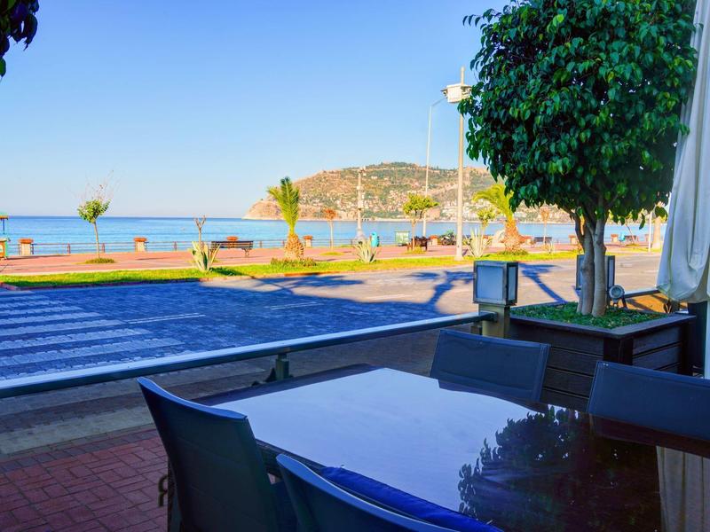 Blick von Terrasse mit Tischen und Stühlen auf eine Straße, Palmen und das Meer im Hintergrund.