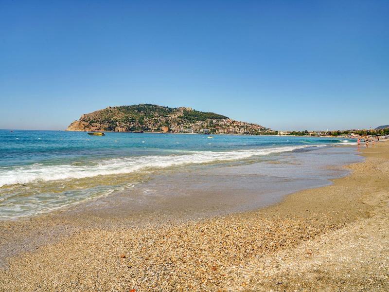 Weitläufiger Sandstrand mit sanften Wellen, klarem Himmel und bewachsenem Hügel im Hintergrund.