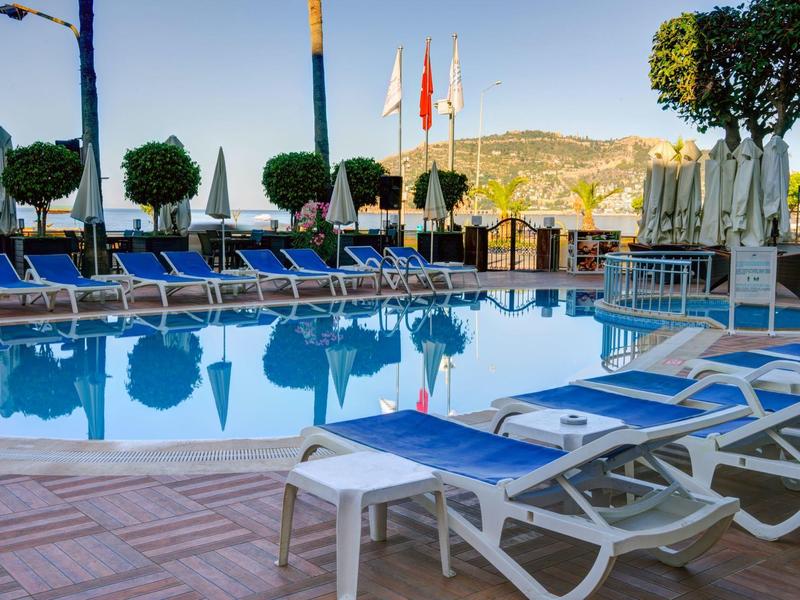 Einladender Pool mit blauen Liegen und weißen Sonnenschirmen vor grünen Bäumen und Bergen im Hintergrund.