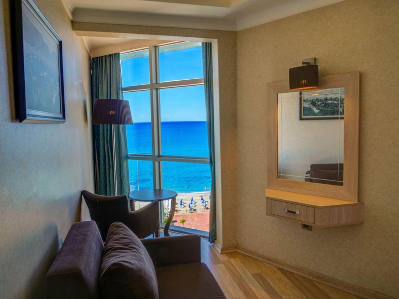 Zimmer mit braunem Sofa, großem Fenster mit Meerblick und Wandspiegel mit Regal.