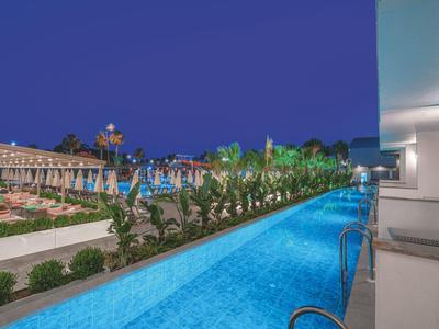 Beleuchteter Pool mit blauem Wasser, Palmen und Abendhimmel, modernes Hotel mit Terrasse rechts.
