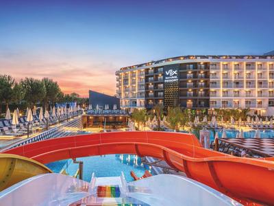 Modernes Hotel mit beleuchtetem Außenpool, Wasserrutsche und Palmen bei Sonnenuntergang.