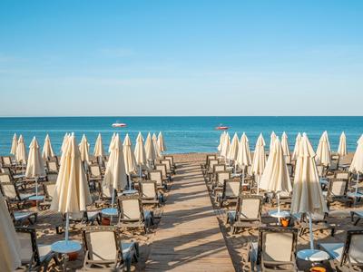 Strand mit vielen weißen Sonnenschirmen und Liegestühlen, blauer Himmel und ruhiges Meer im Hintergrund.