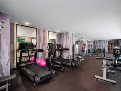 Moderner Fitnessraum mit Laufbändern, Hanteln, Trainingsgeräten und lila Vorhängen an den Fenstern.