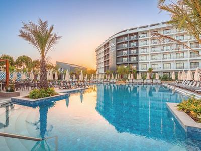 Großer Pool mit blauer Wasserfläche, Palmen und modernen Hotelgebäuden im Abendlicht.