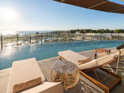 Infinity zwembad met ligstoelen met uitzicht op zee en stad bij zonsondergang.